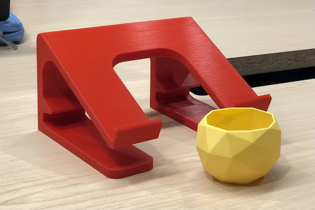 To the left, a red 3D print of a laptop stand, and to the right, a 3D print of a yellow flower pot.