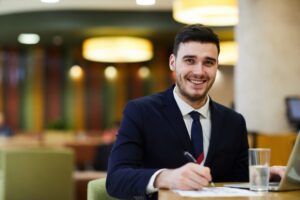 Male hotel apprentice working with a pen, paper and laptop, wearing a blue suit