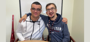 two men in their mid 20s sitting arm and arm smiling in an office.