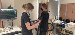 two female students in scrubs. One using a blood pressure cuff on the other to check blood pressure