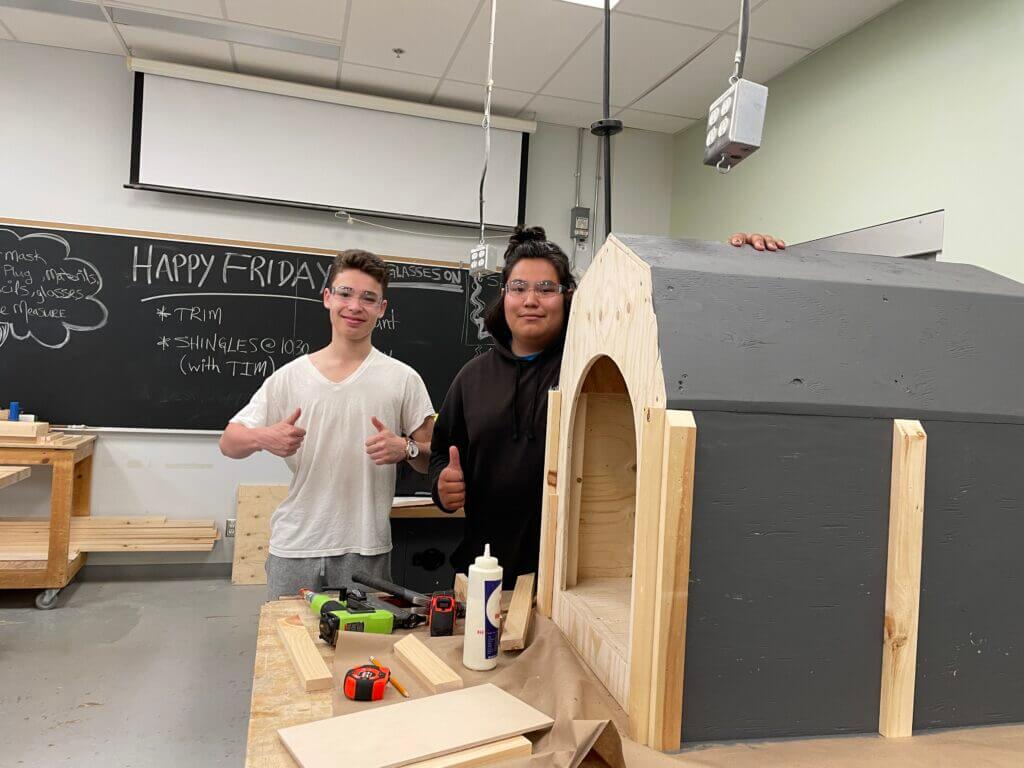 Two students stand to the left of a doghouse construction project; one student is holding two thumbs up.