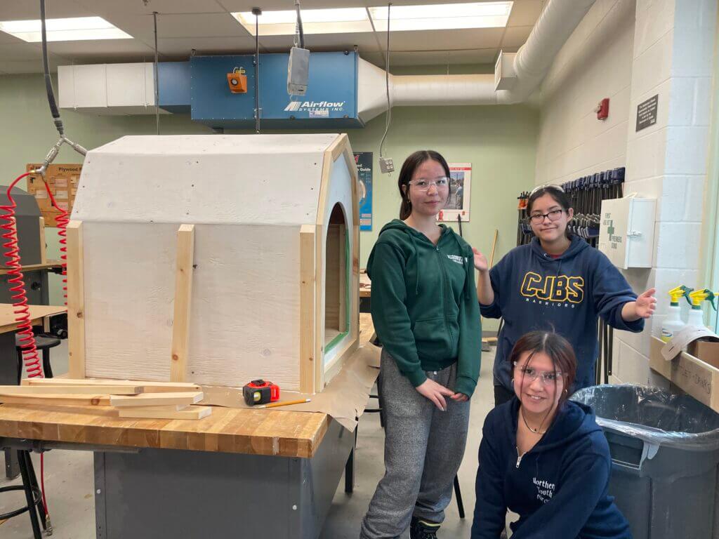 Three students stand to the right of a doghouse construction project; one student is kneeling in front.