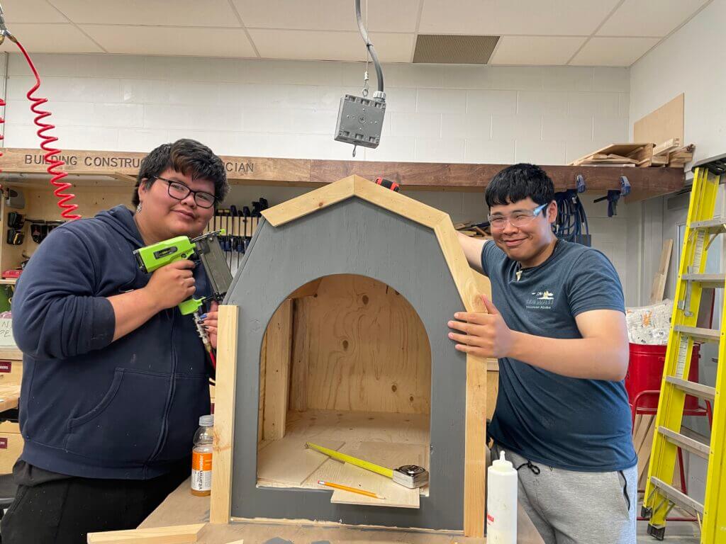 Two carpentry students stand on either side of a doghouse construction project.