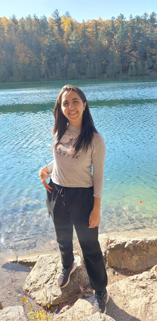 A woman smiles in front of a lakeside forest.