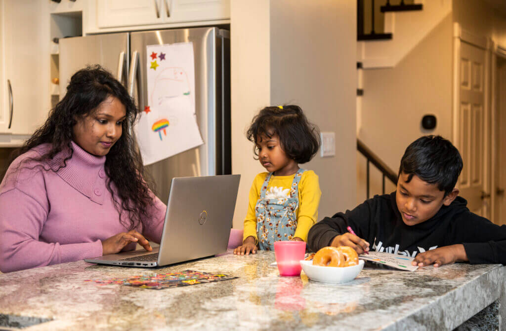 An AC Online mature student works on her laptop at her kitchen island while her two children colour a book