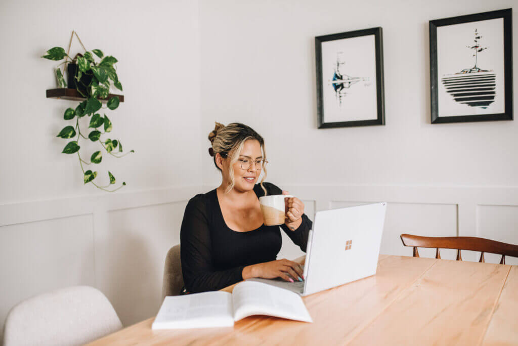 An Indigenous female sits at a table in front of her laptop while holding a coffee mug. An open textbook sits to her right.