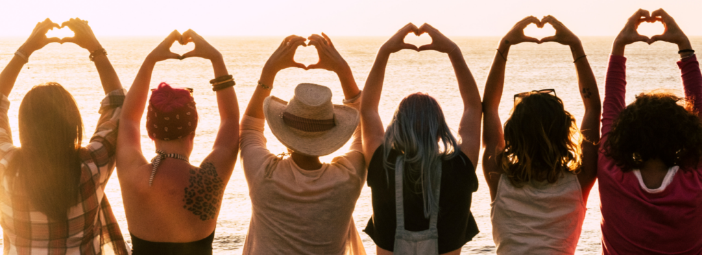 Group of women facing a beach sunset with their hands all making heart signs