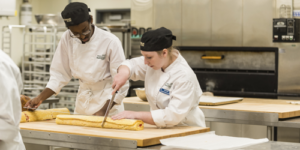 Female student cutting a pastry at the culinary lab with her classmate next to her.