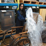 Student removing plastic sheet covering an old wooden wheelchair in a warehouse