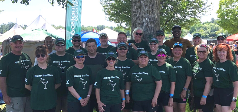 The Bizzy Barflies team stands together outside with trees in the background. All team members where forest green shirts with a graphic that reads "Bizzy Barflies" with a cartoon martini glass and buzzing fly. 