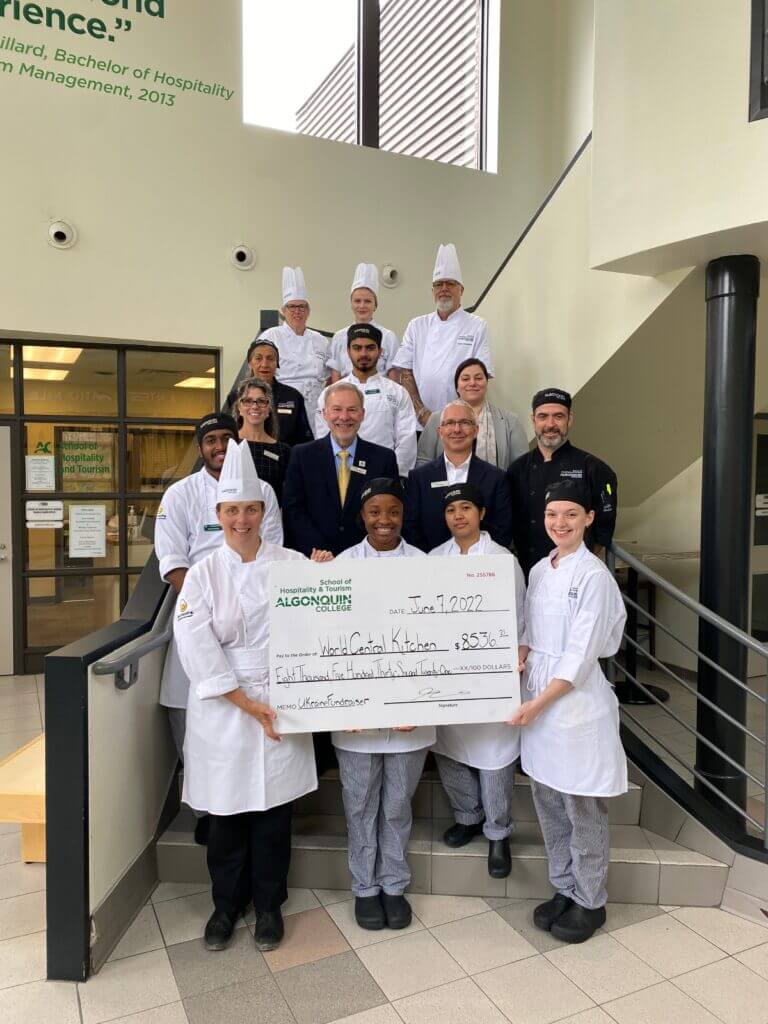 Chef Skeates (bottom left) and President &amp; CEO Claude Brulé (second row from bottom, middle-left) are joined by students and faculty who were integral to the fundraiser's success. The pictured individuals stand staggered on the stairs with the front four people holding up a large check. 