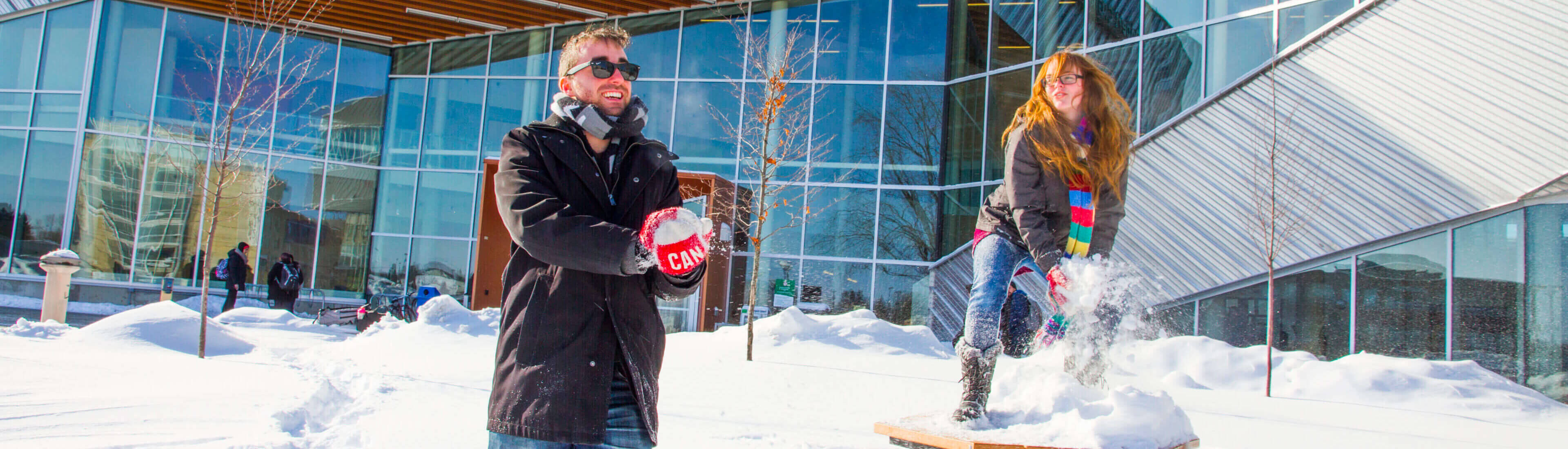 Students throwing snow at each other in front of the student commons on a sunny day in the winter. The campus is covered in snow.