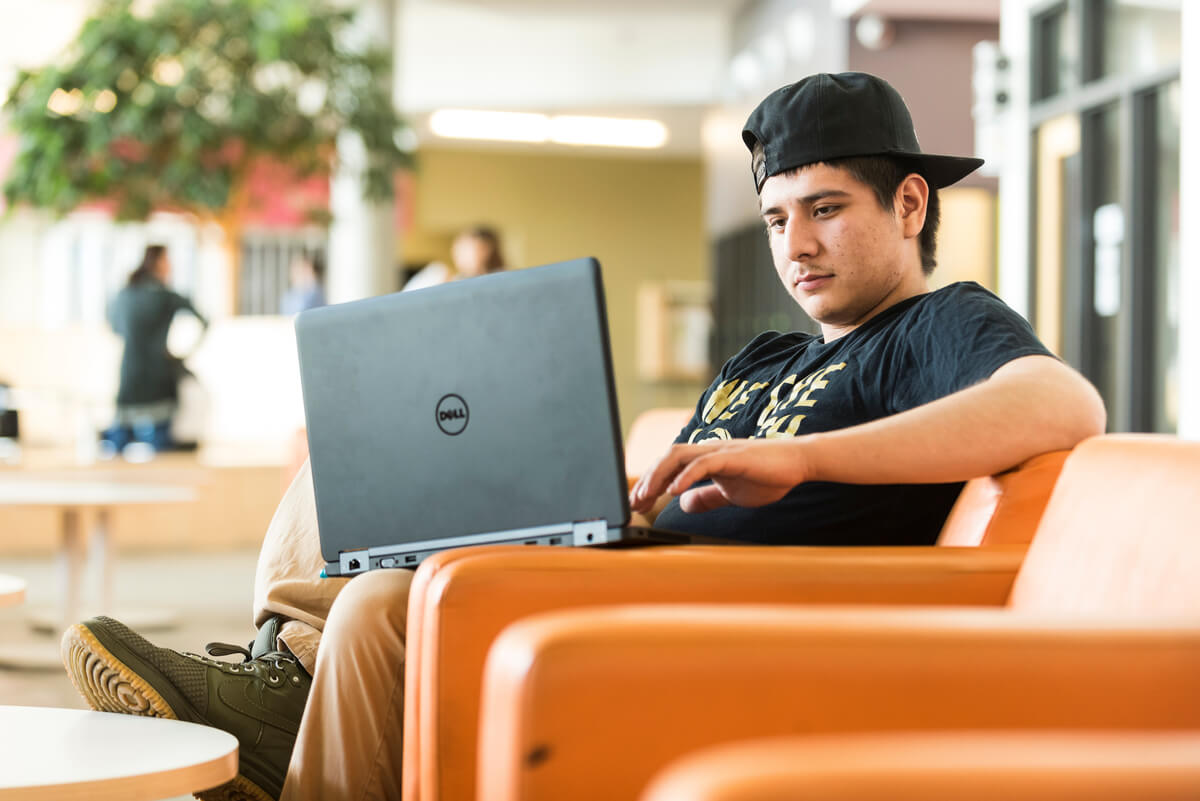 An Algonquin College student is sitting in a chair and using a laptop computer.