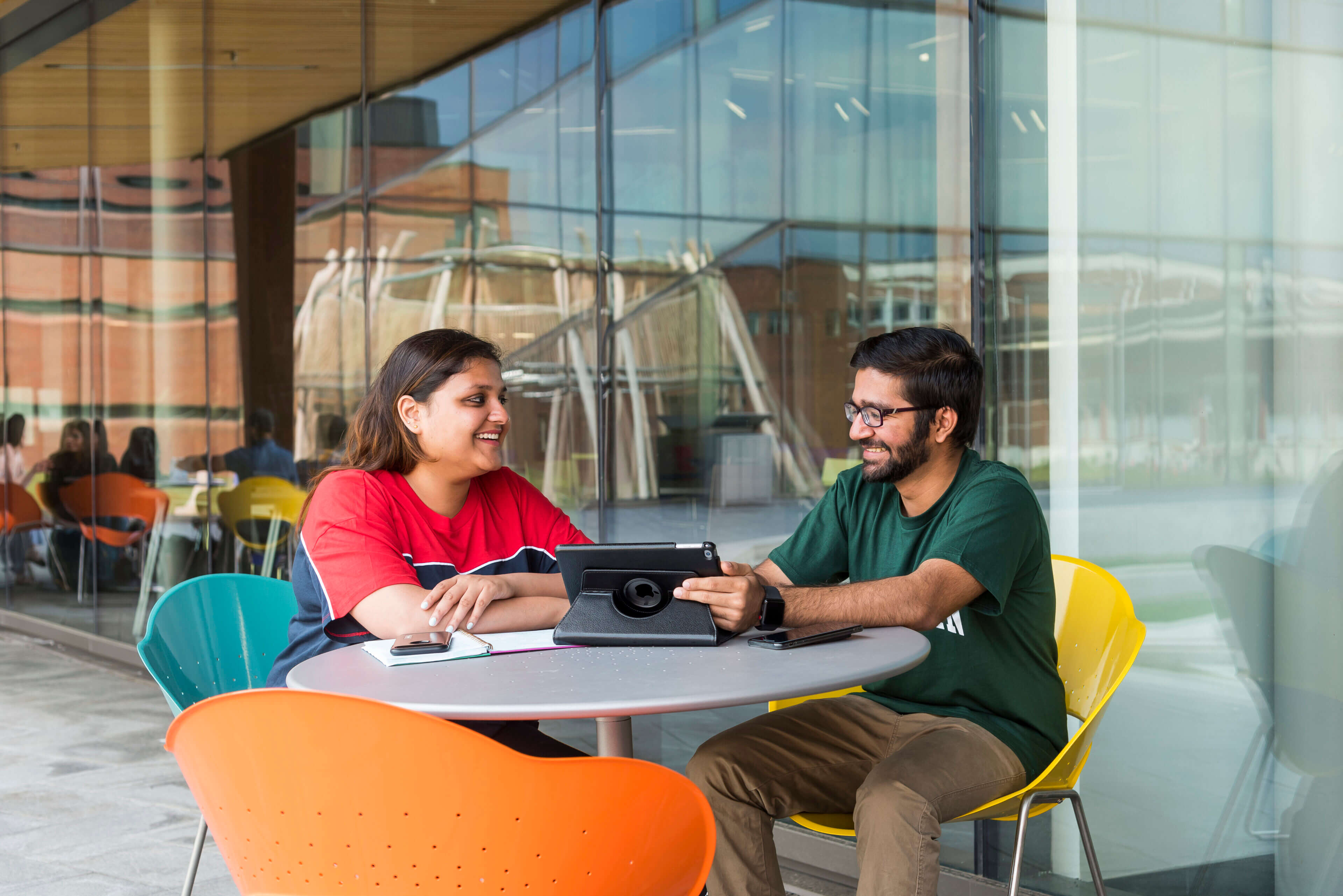 A man and a woman are smiling and sitting at a table at the Ishkodewan courtyard while the man holds a tablet.