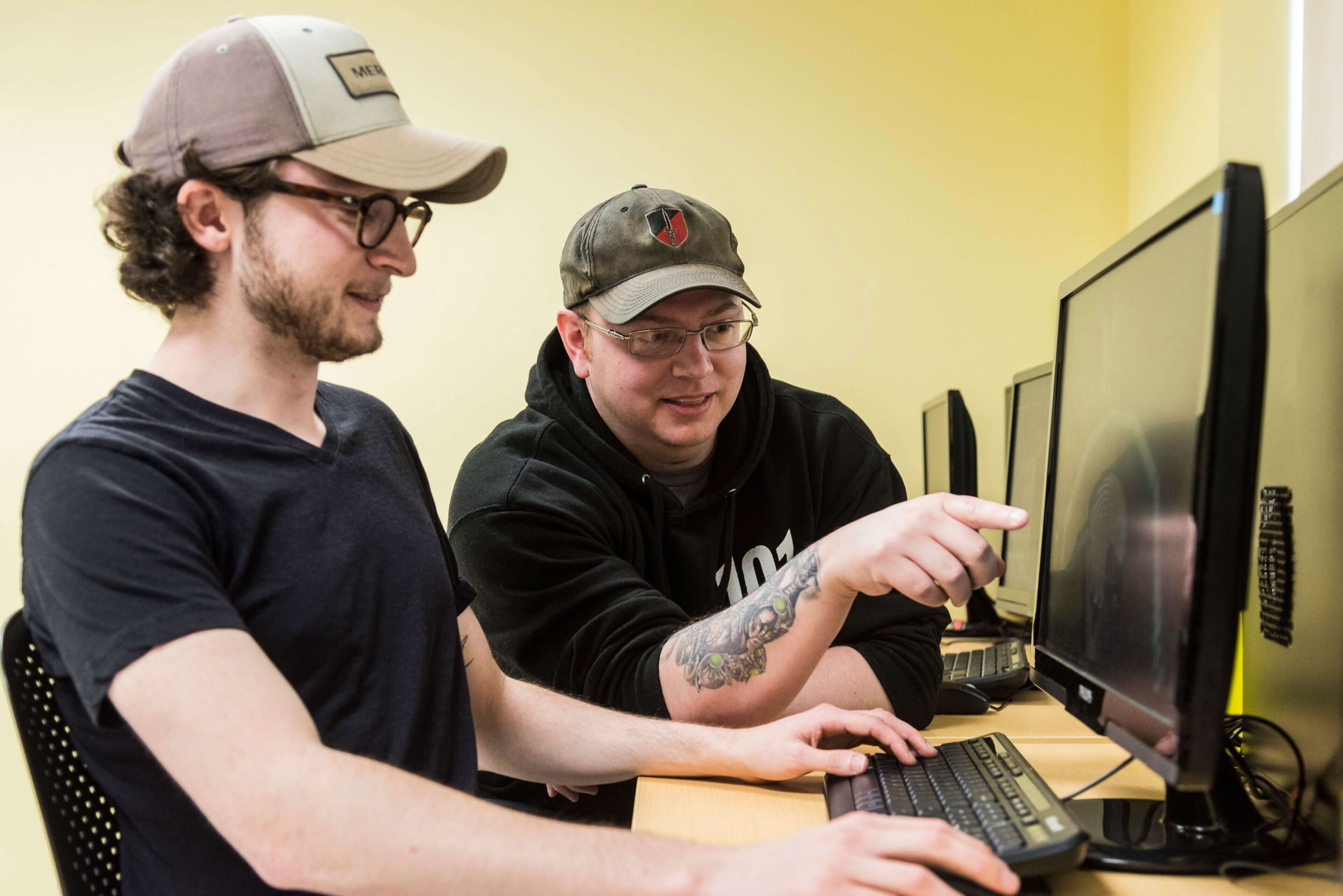 Two young men are sitting at a desk. The first one is facing a computer screen while he holds a mouse and rests his left hand on the keyboard. The second one is beside him, leaning forward and pointing at something on the screen.