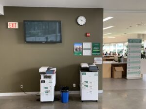 An image of two different Ricoh multi function printers located on the second floor of E building, in the hallway outside of the AC Hub.