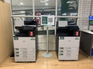 An image of two Ricoh multi function printers in the Student Print Shop located within Connections: The Campus Store room H110.